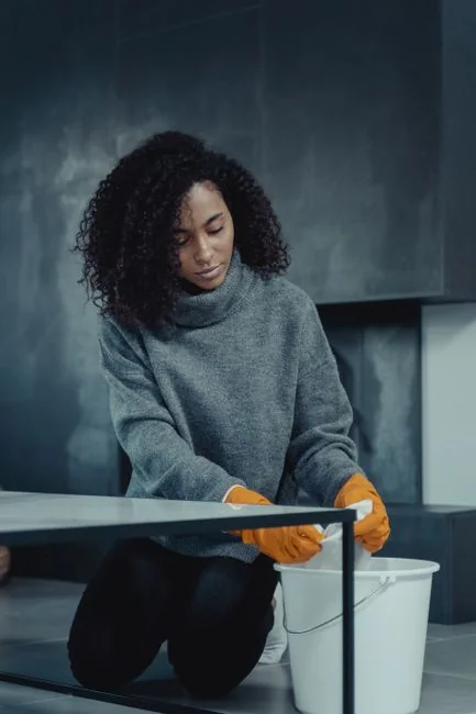 Side view of young focused female in latex gloves standing and cleaning mirror in light room in daytime
